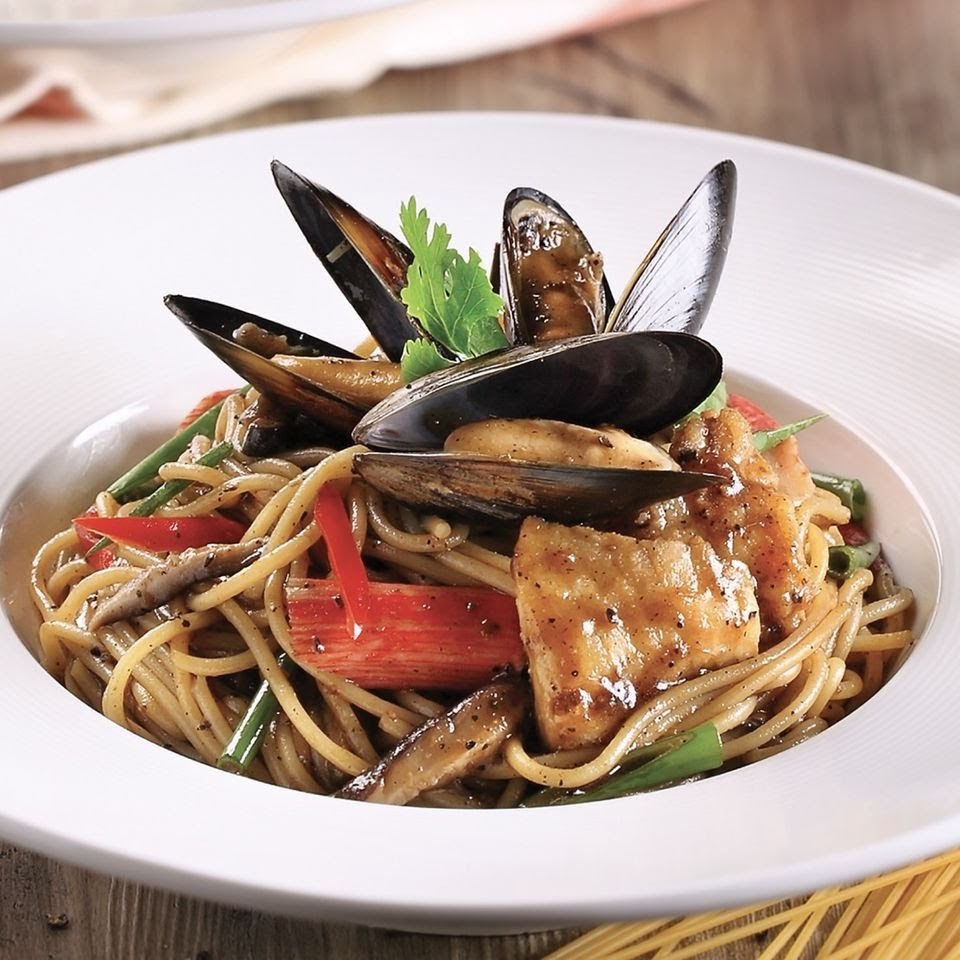 A close-up of a delicious seafood spaghetti dish with mussels, fish, and vegetables in a white bowl.