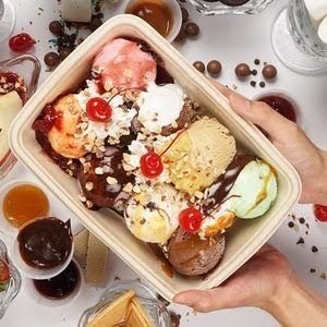 A large takeaway container of colorful ice cream scoops with toppings, held by hands.