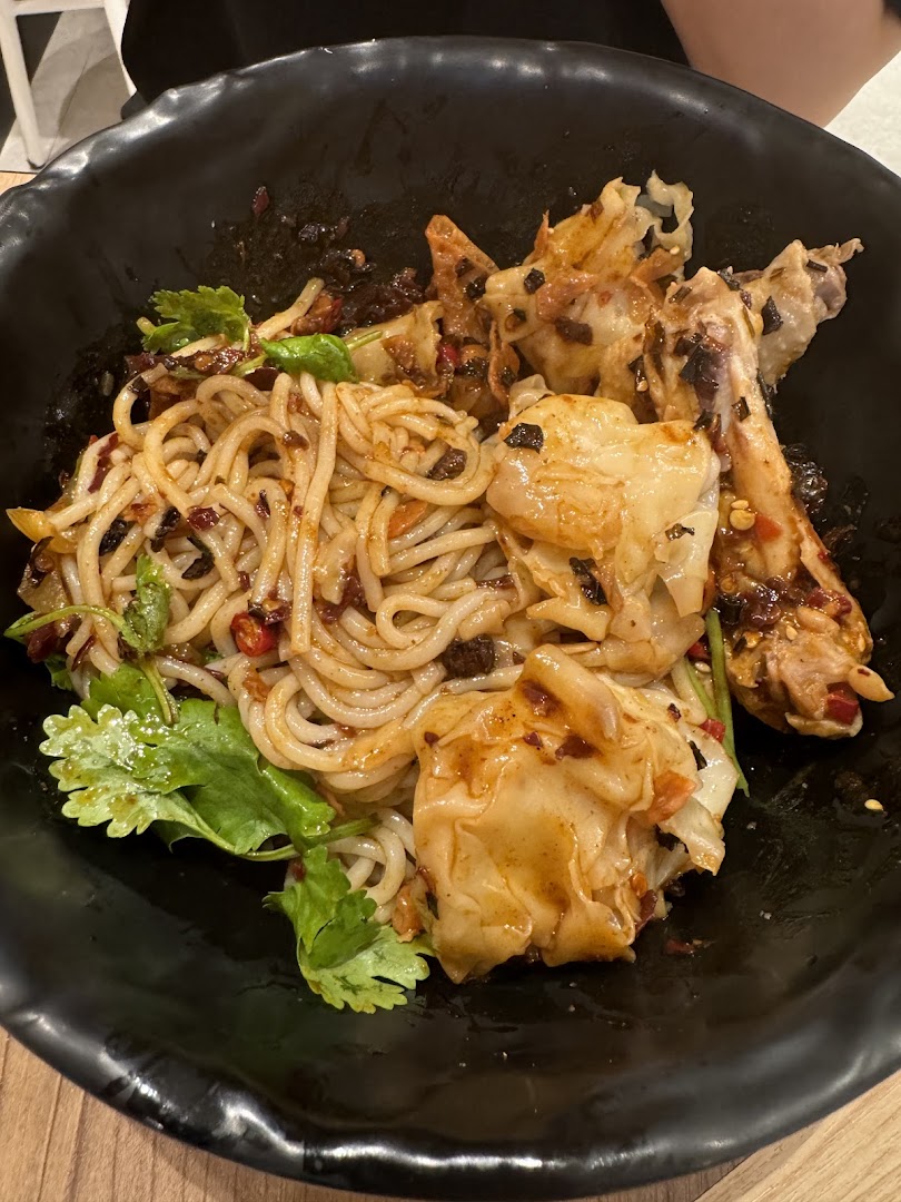 A close-up of a delicious bowl of spicy noodles with dumplings and chicken in a black bowl.