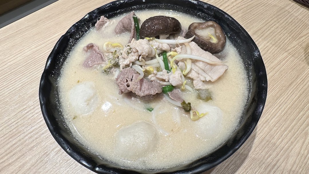 A close-up of a bowl of TamJai SamGor Mixian with meat, mushrooms, and fish balls in a creamy broth.