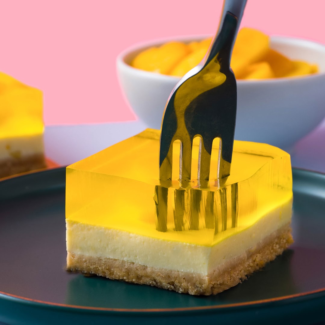 A fork pierces a vibrant yellow jelly dessert with creamy and crumbly layers on a dark plate.
