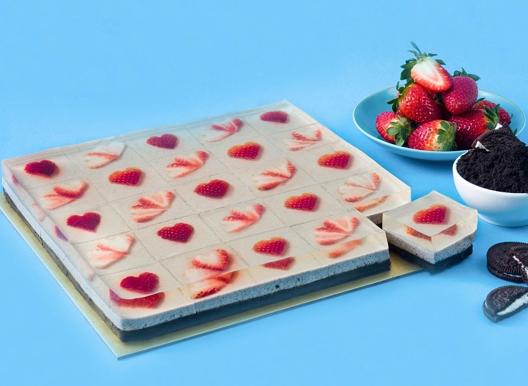 A vibrant jelly cake with strawberry hearts and slices, alongside fresh strawberries and crushed cookies.