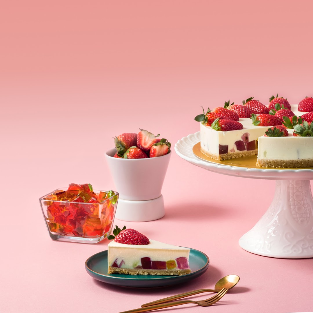 A vibrant image showcasing a cheesecake with colorful jelly cubes and fresh strawberries on a pink background.
