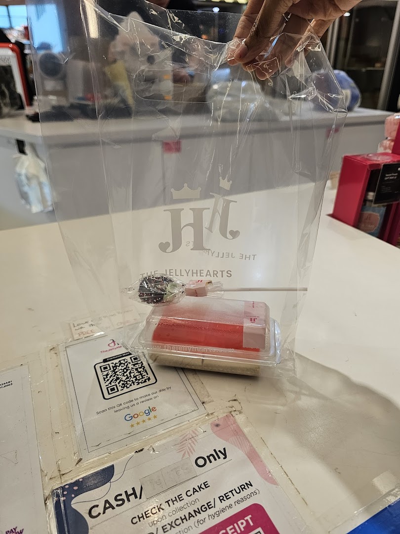 A hand holds a clear bag with The JellyHearts logo, containing a cake pop and a jelly dessert on a counter.