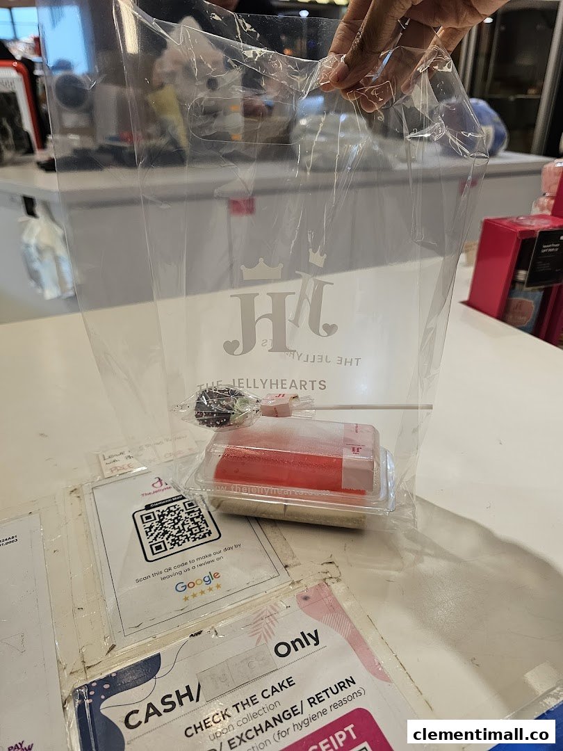 A hand holds a clear bag with The JellyHearts logo, containing a cake pop and a jelly dessert on a counter.