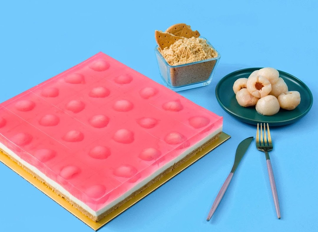 A vibrant pink jelly cake with lychee pieces, served with crushed biscuits and fresh lychees.