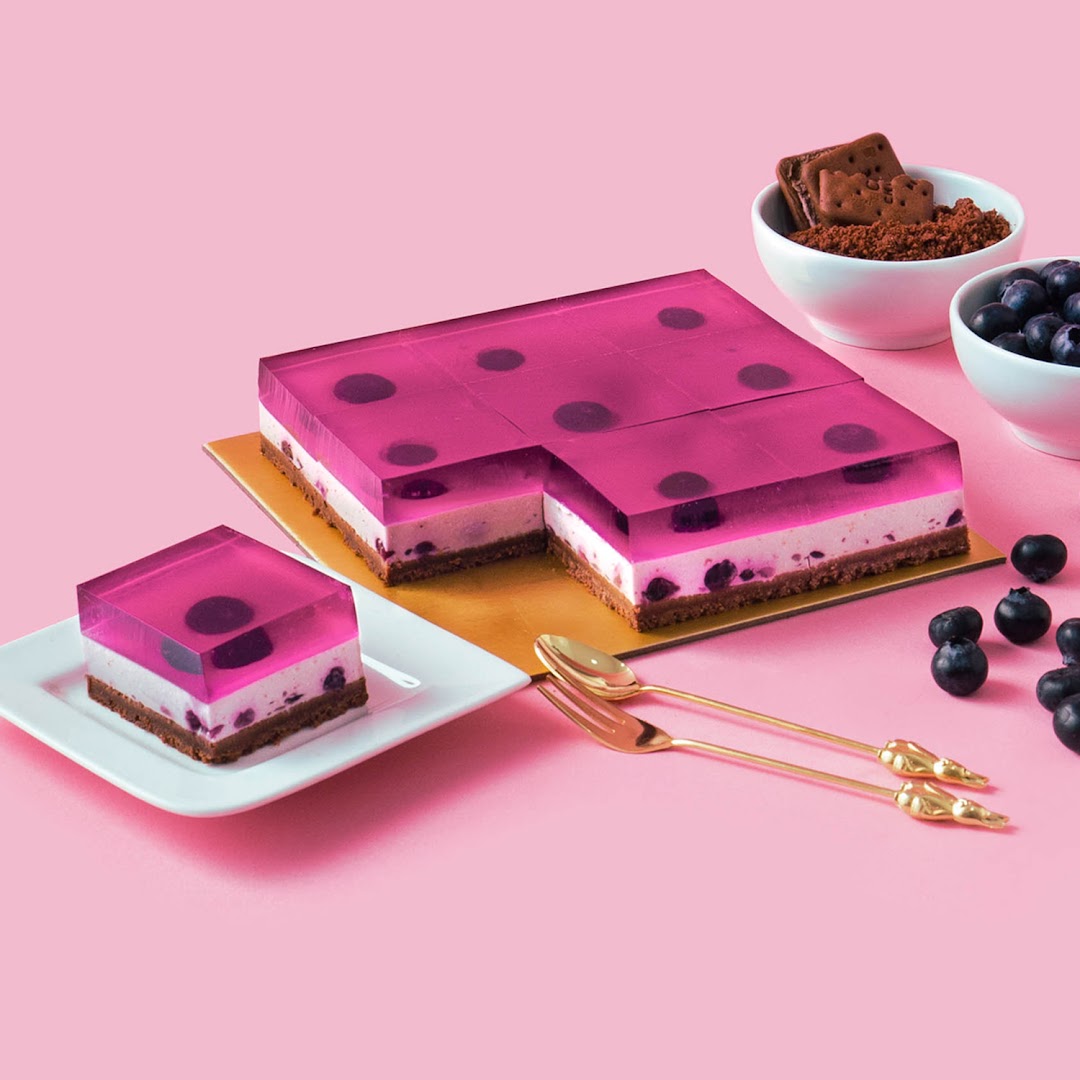 A vibrant blueberry jelly cheesecake with a slice served on a plate, set against a pink background.