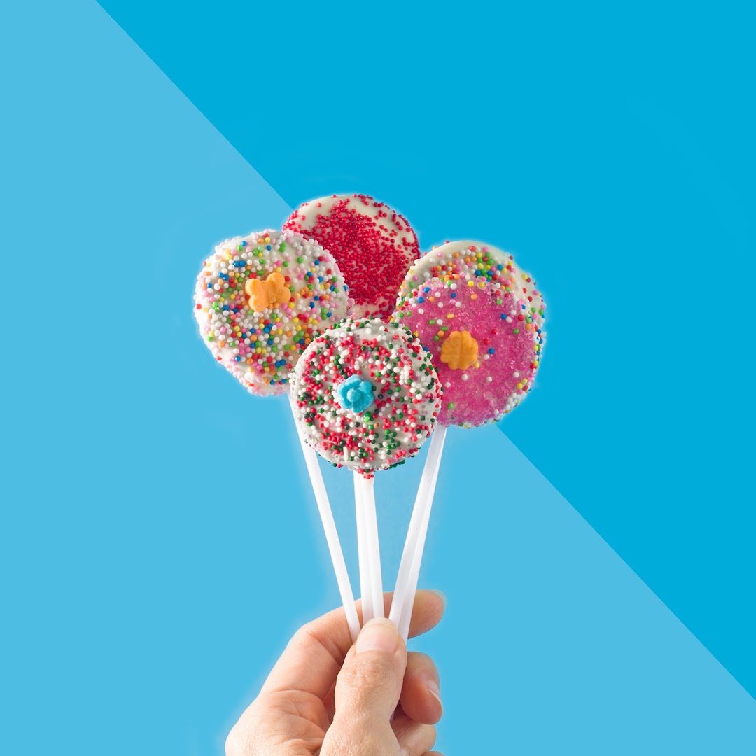 A hand holds four colorful, sprinkle-covered lollipops against a vibrant blue background.