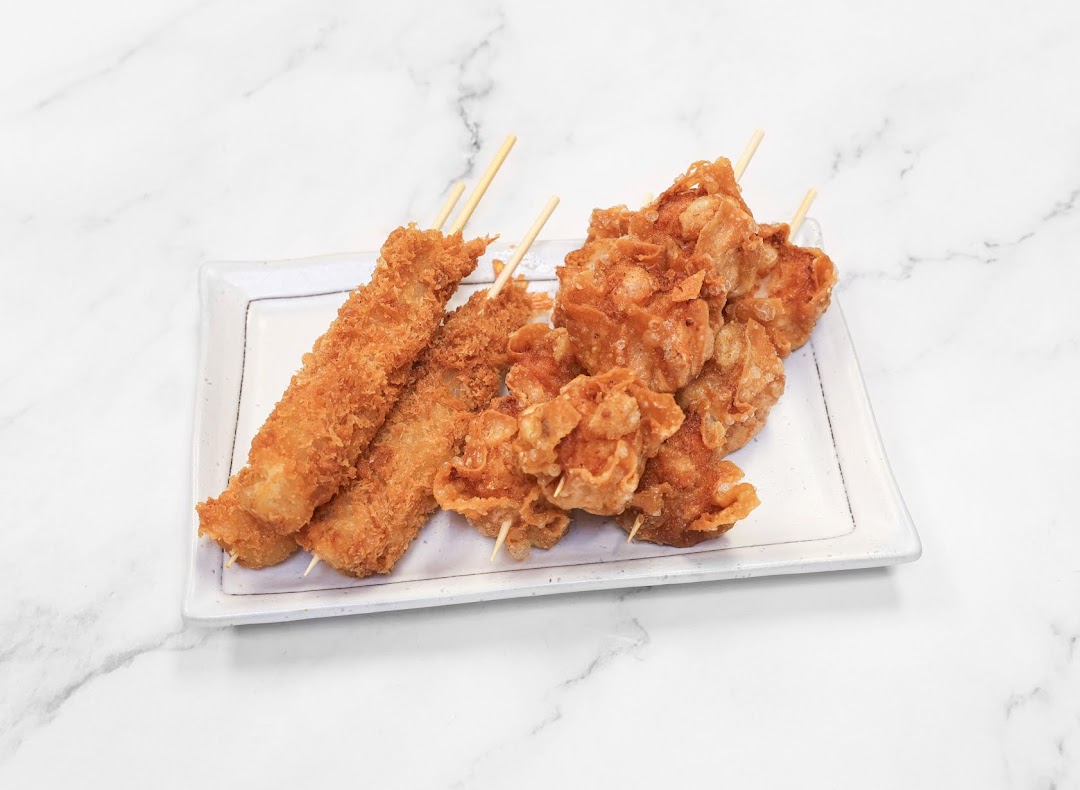 A plate of crispy fried skewers, including breaded and cornflake-crusted varieties, on a white marble background.