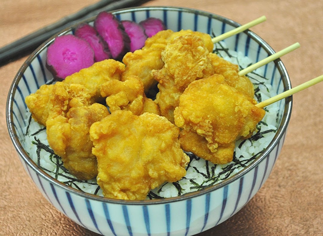 A bowl of golden fried chicken pieces on rice with pickled vegetables and chopsticks.