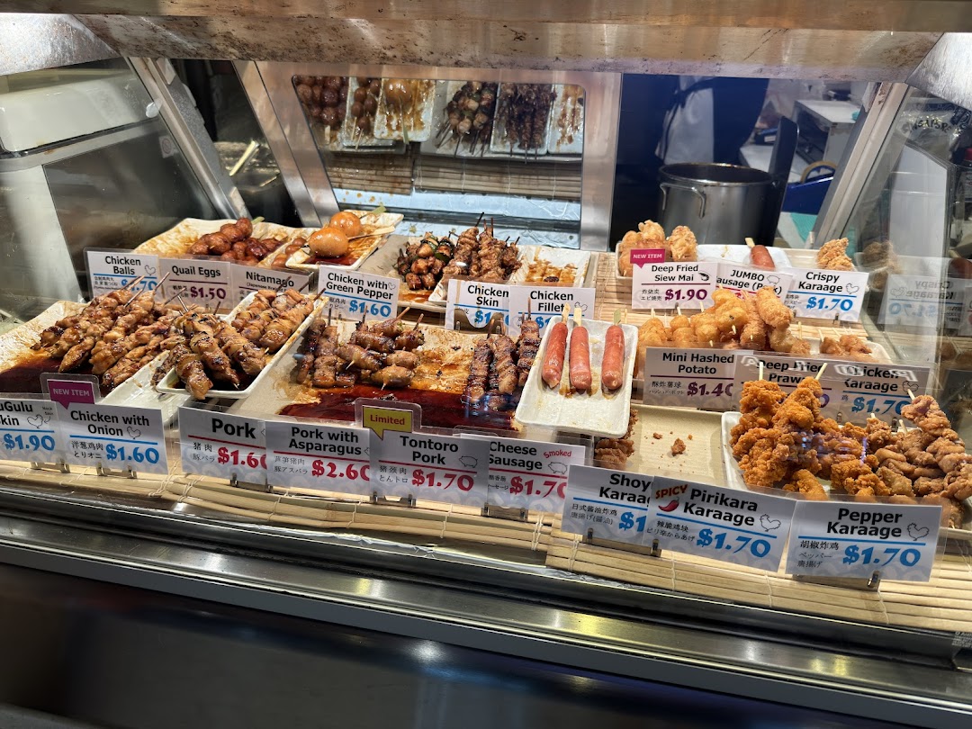 A vibrant display case showcasing a variety of Japanese skewers, fried items, and snacks with prices.