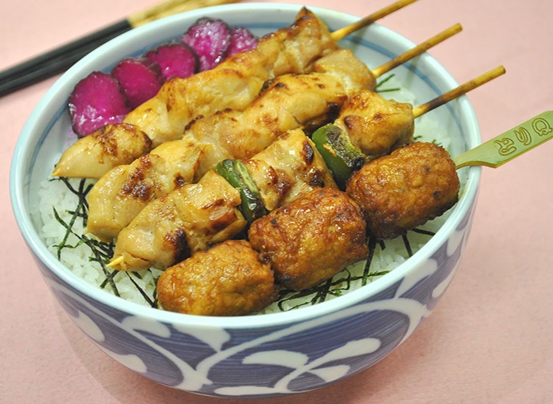 A vibrant bowl of Japanese yakitori skewers and meatballs served over rice with pickled vegetables.