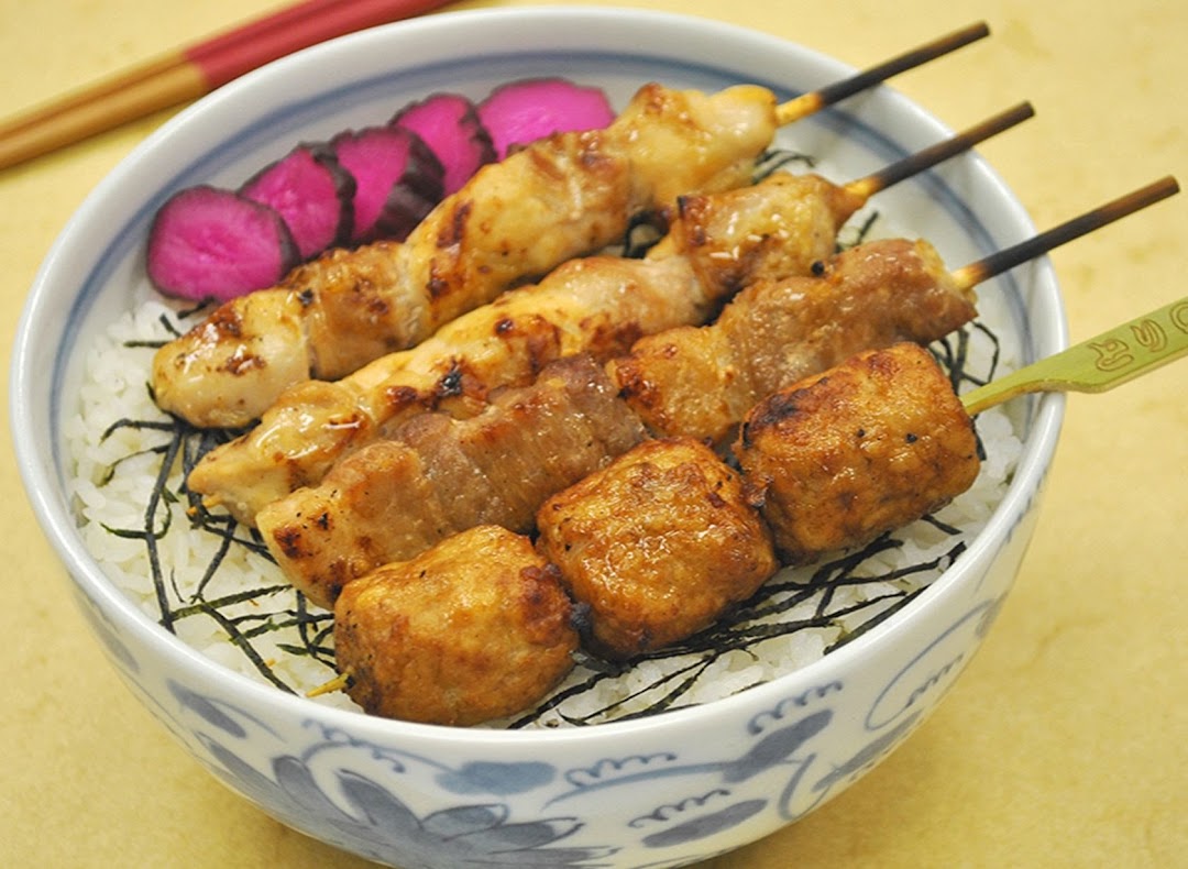 A bowl of rice topped with various grilled yakitori skewers and pickled vegetables.