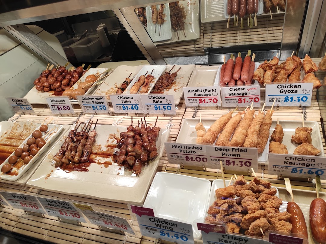 A vibrant display counter showcasing various Japanese skewers, fried items, and sausages with prices.