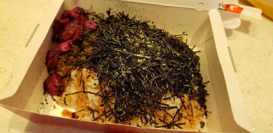 A close-up of a Japanese rice bowl topped with shredded seaweed and pickled vegetables in a takeaway box.