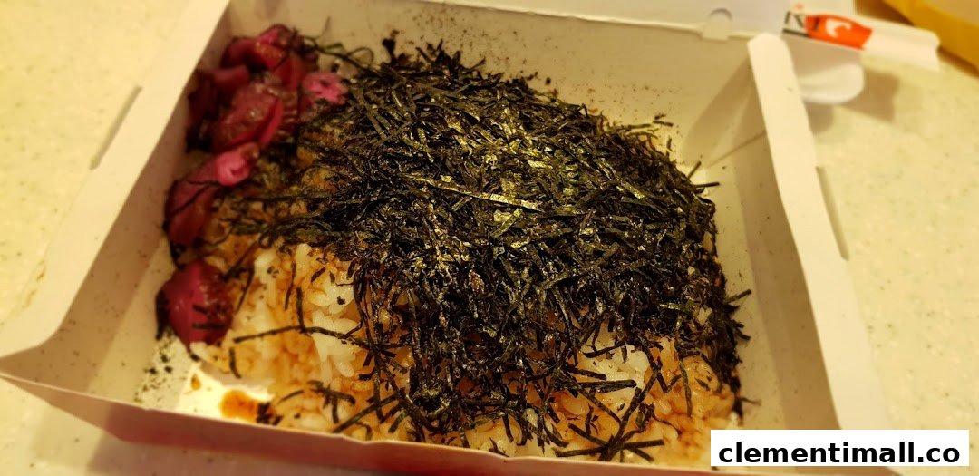A close-up of a Japanese rice bowl topped with shredded seaweed and pickled vegetables in a takeaway box.