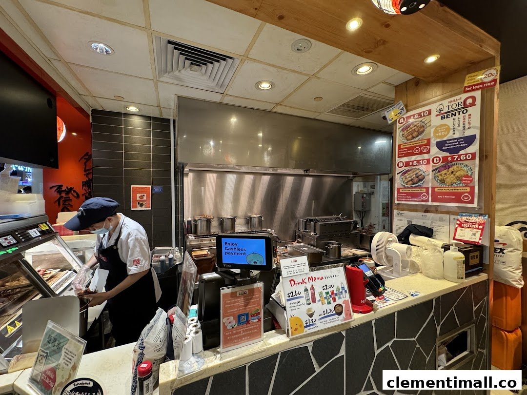 A staff member prepares food at the counter of TORI-Q, with menu boards and payment options visible.