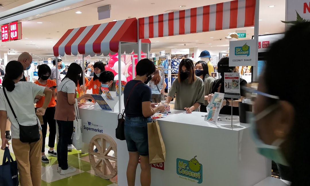 Shoppers interact with staff at a vibrant Mr. Coconut pop-up stall inside UNIQLO, featuring Shopee Supermarket branding.