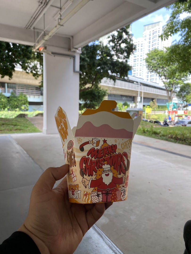 A hand holds a festive Wok Hey noodle box outdoors, featuring a Christmas-themed design.