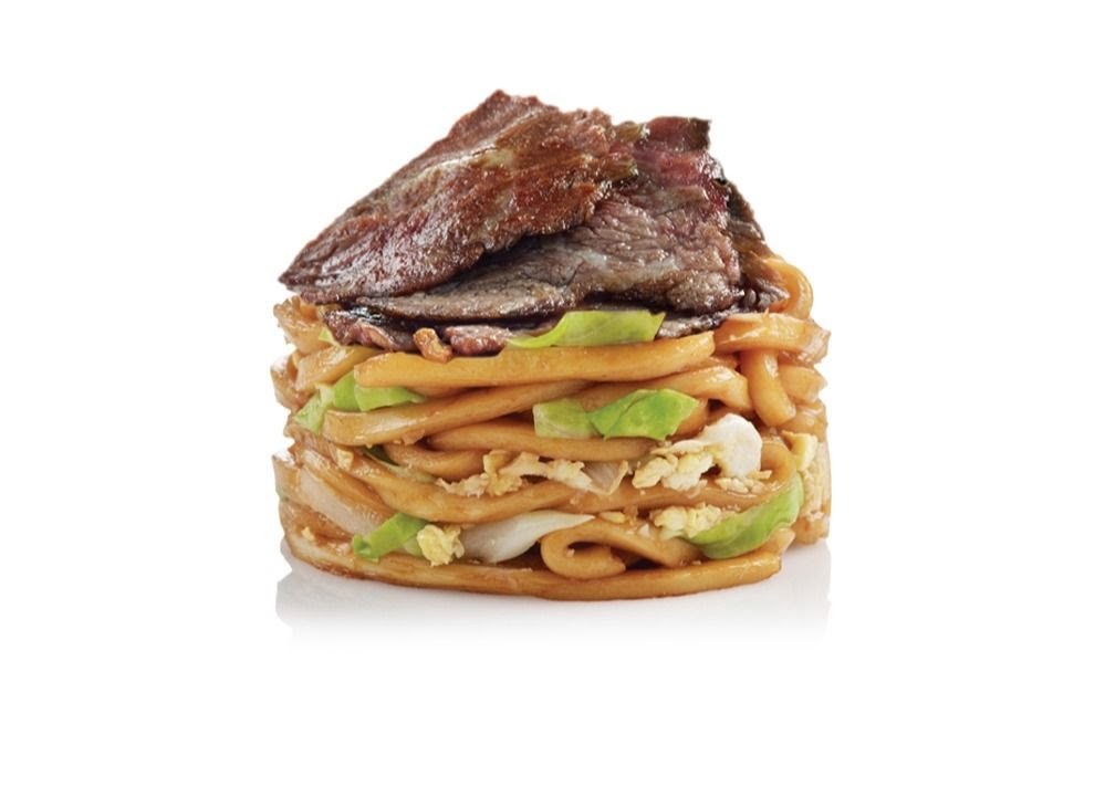 A close-up of a stacked noodle dish topped with sliced beef and vegetables.