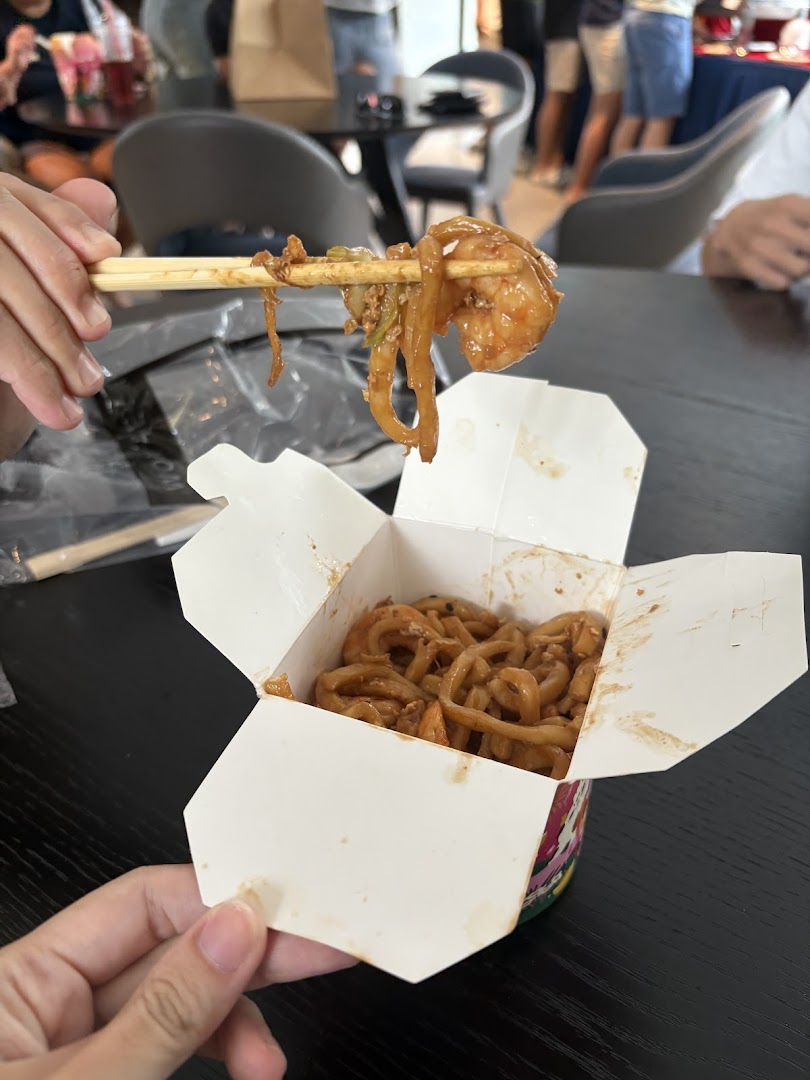 A person holds chopsticks with noodles and shrimp from a WOK HEY take-out box.