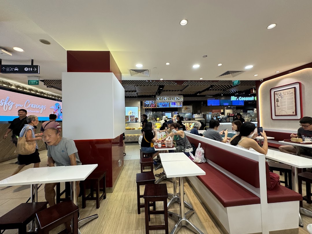 An interior view of Ya Kun Kaya Toast, showing customers seated at tables and benches in a bustling food court setting.