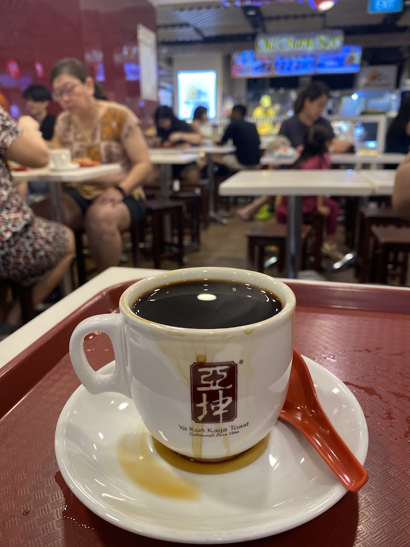 Close-up of a signature coffee cup from Ya Kun Kaya Toast on a tray, with blurred diners in the background.