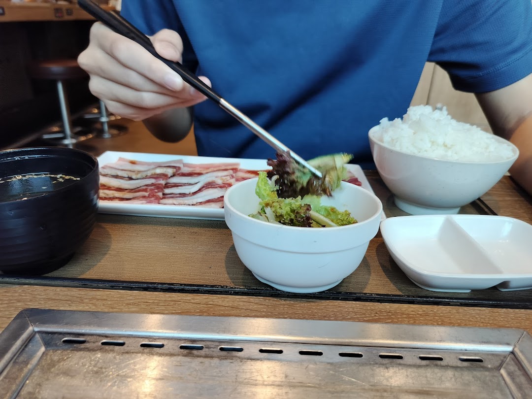 A diner uses chopsticks to pick up salad, with raw meat, rice, and a grill on the table.
