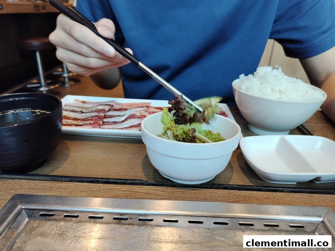 A diner uses chopsticks to pick up salad, with raw meat, rice, and a grill on the table.