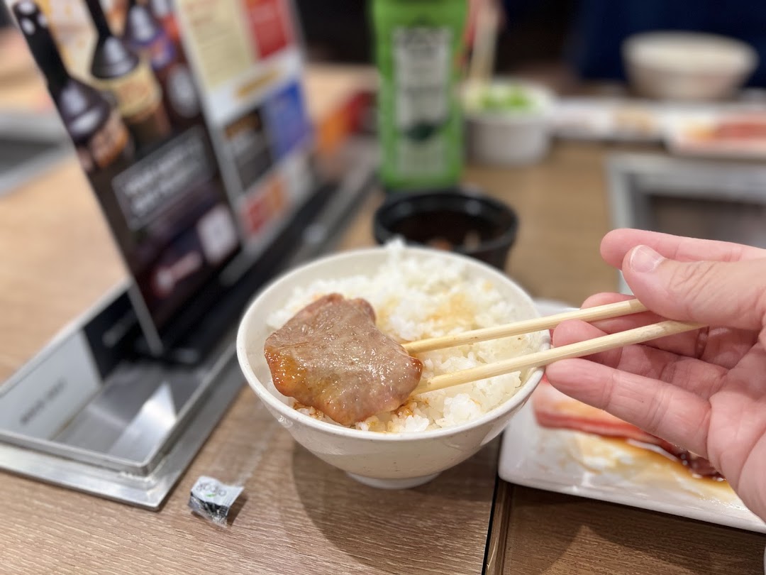 A hand with chopsticks picks up a piece of grilled meat from a bowl of rice at a yakiniku restaurant.