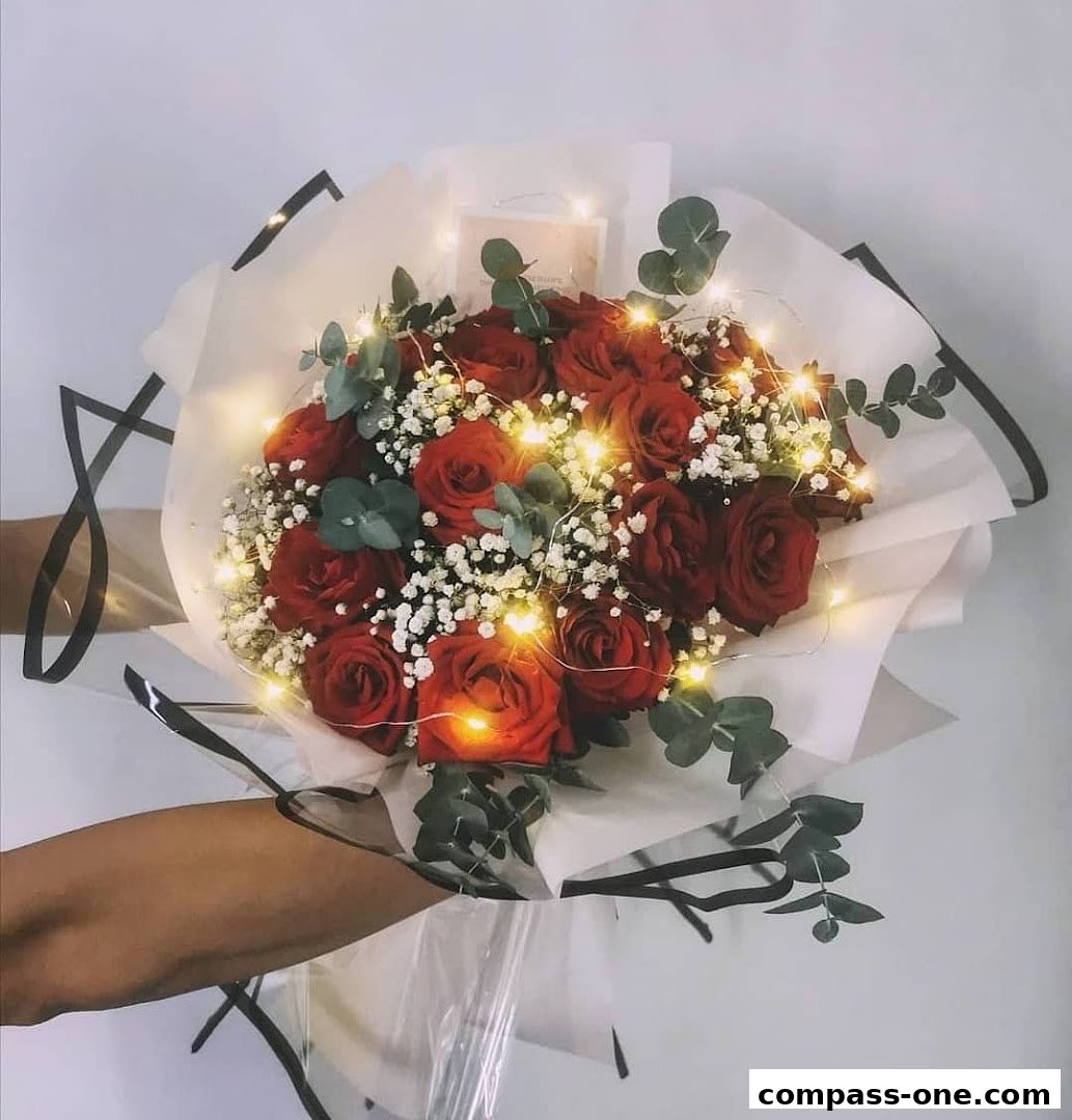 A hand holding a bouquet of red roses with baby's breath and eucalyptus, adorned with fairy lights.