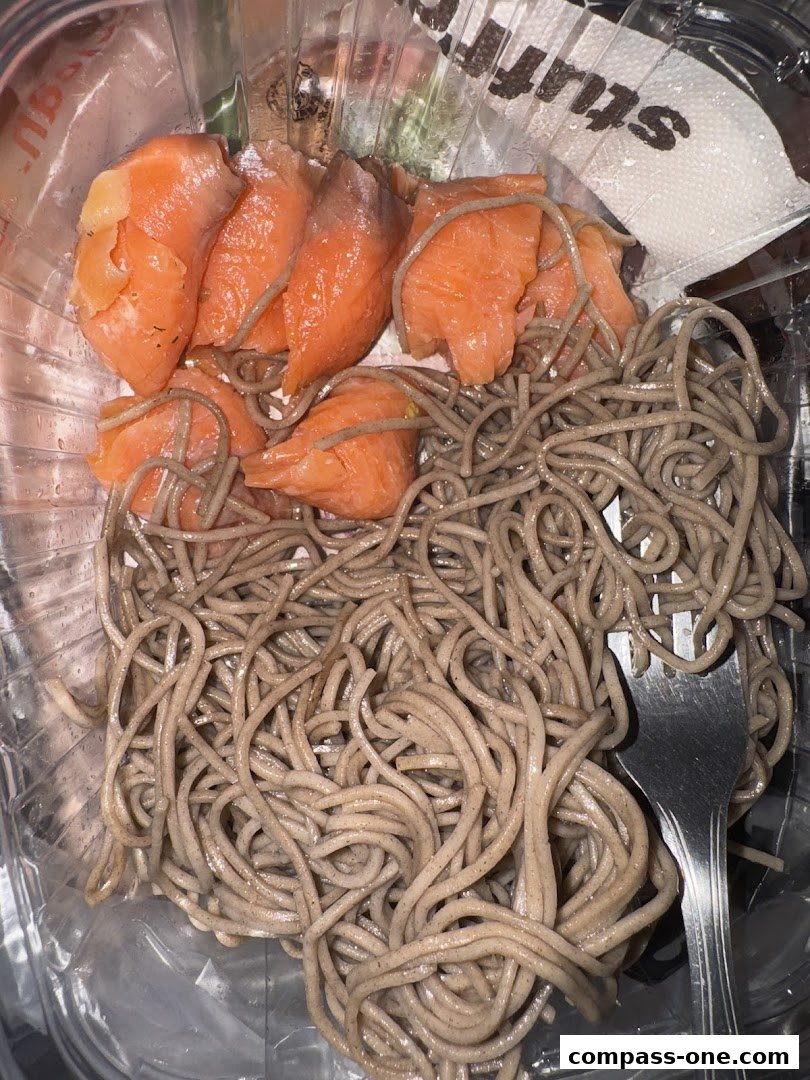 A container of soba noodles topped with slices of smoked salmon and a fork.