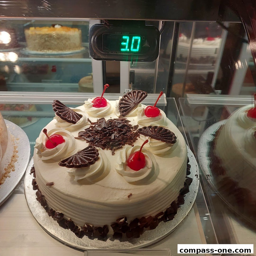 A beautifully decorated white cake with cherries and chocolate shavings sits in a display case.