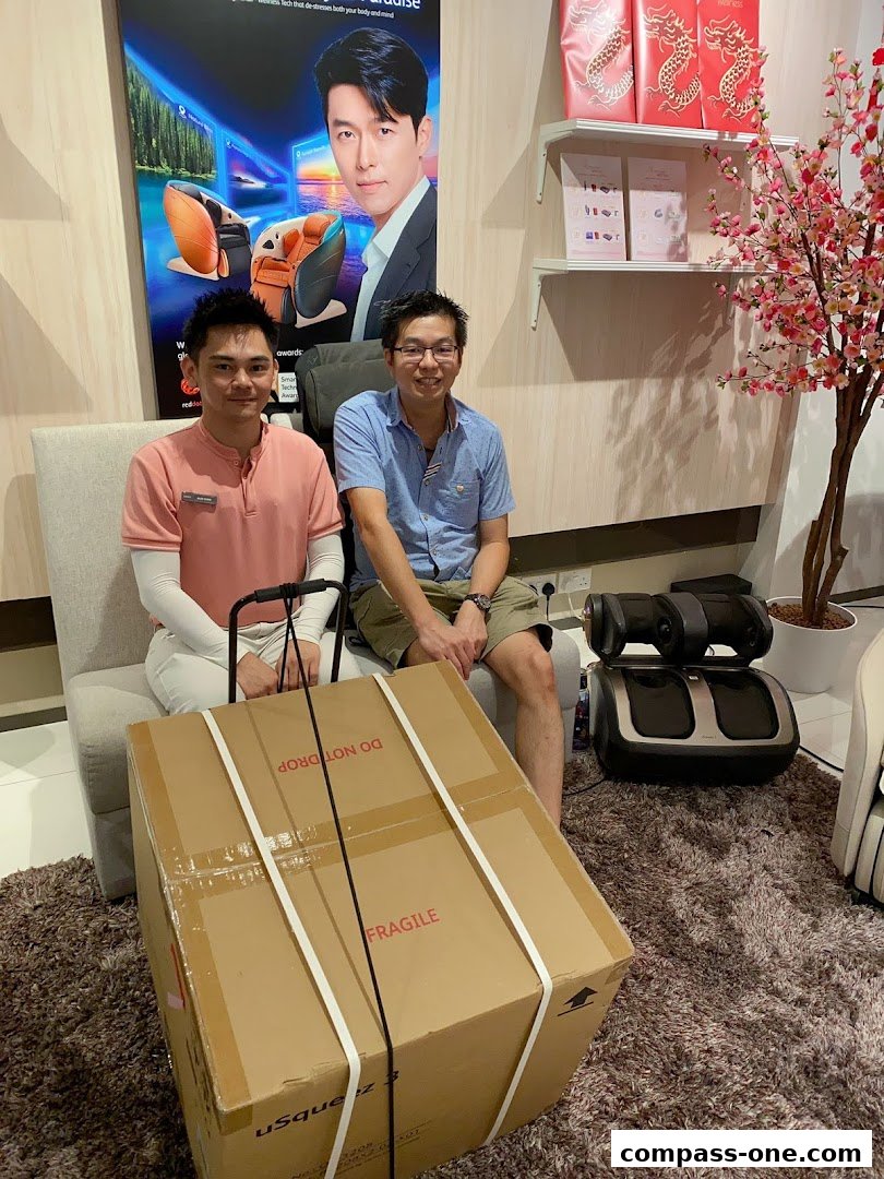 Two men are posing with a large boxed product in a retail store, with massage chairs and decor in the background.