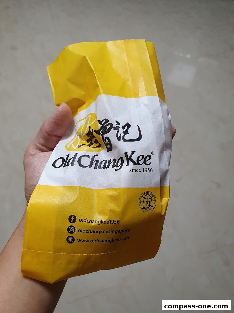 A hand holds a yellow and white paper bag with the Old Chang Kee logo and social media handles.