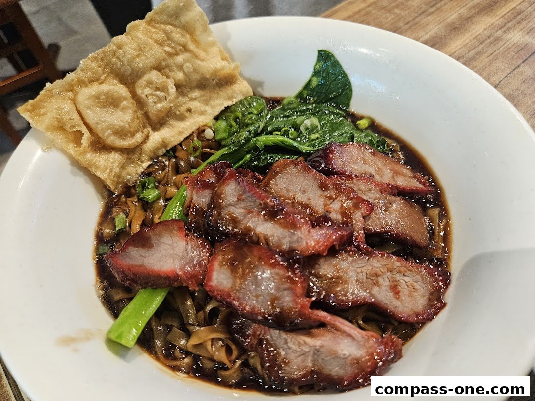 A bowl of Ipoh-style noodles with char siu pork, greens, and a crispy wonton.