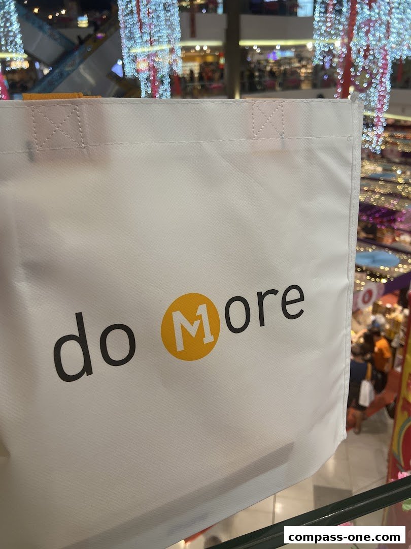 A white shopping bag with 'do M1 More' logo against a blurred mall background with festive lights.