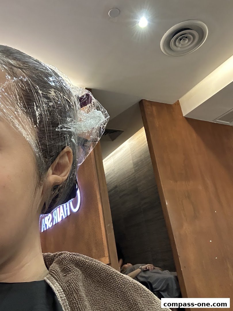 A person's head is covered in plastic wrap, with a towel around their shoulders, inside a salon.