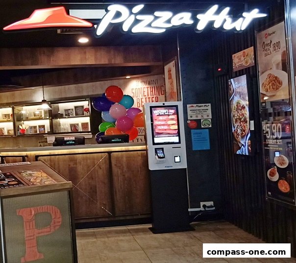The entrance of a Pizza Hut restaurant with a glowing sign, balloons, and a digital ordering kiosk.