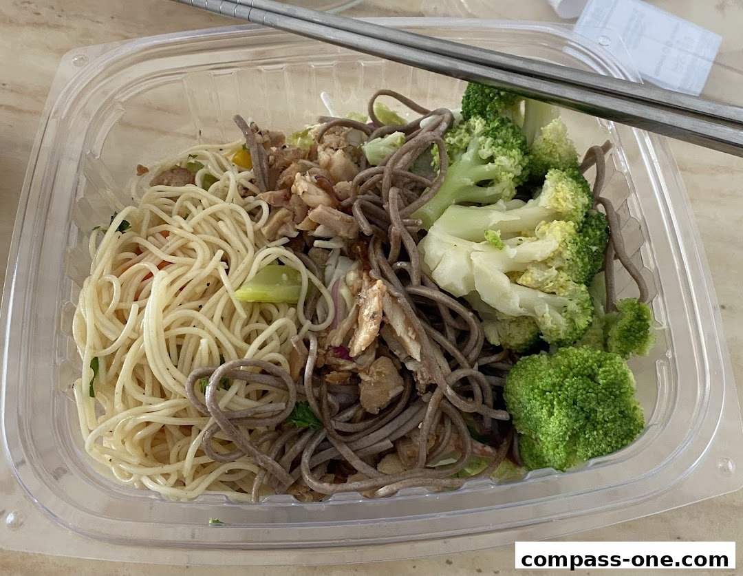 A clear plastic container filled with two types of noodles, chicken, and broccoli, with chopsticks resting on top.
