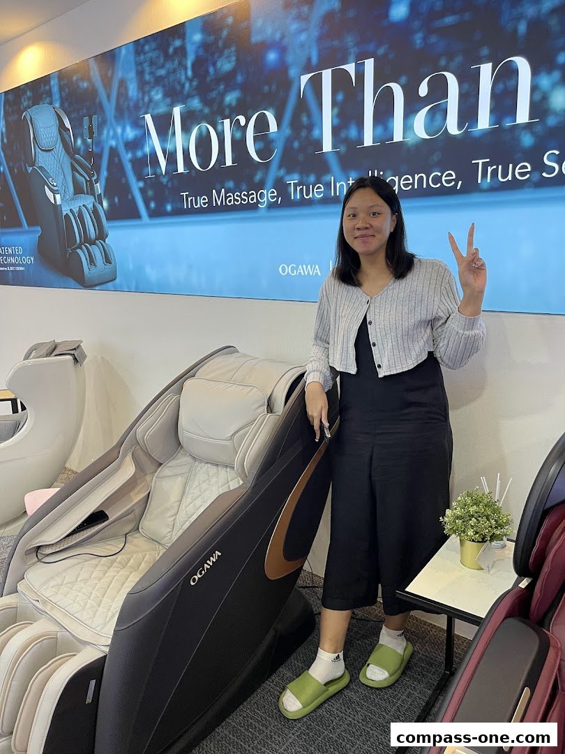 A woman poses with a massage chair in a retail store, with a large advertisement in the background.
