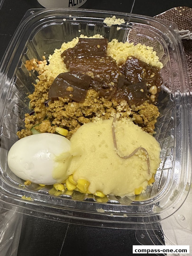 A clear plastic container filled with a meal featuring couscous, meat, mashed potatoes, and a hard-boiled egg.