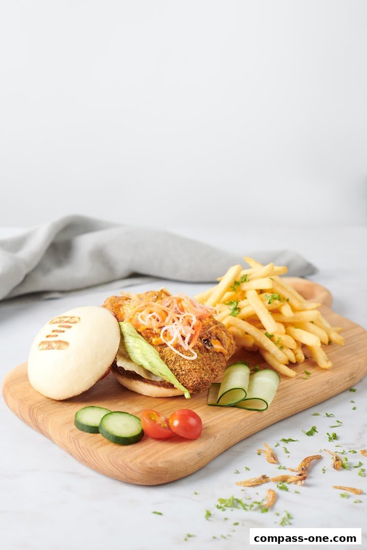 A delicious-looking bao bun burger with crispy fries, cucumber slices, and cherry tomatoes on a wooden board.