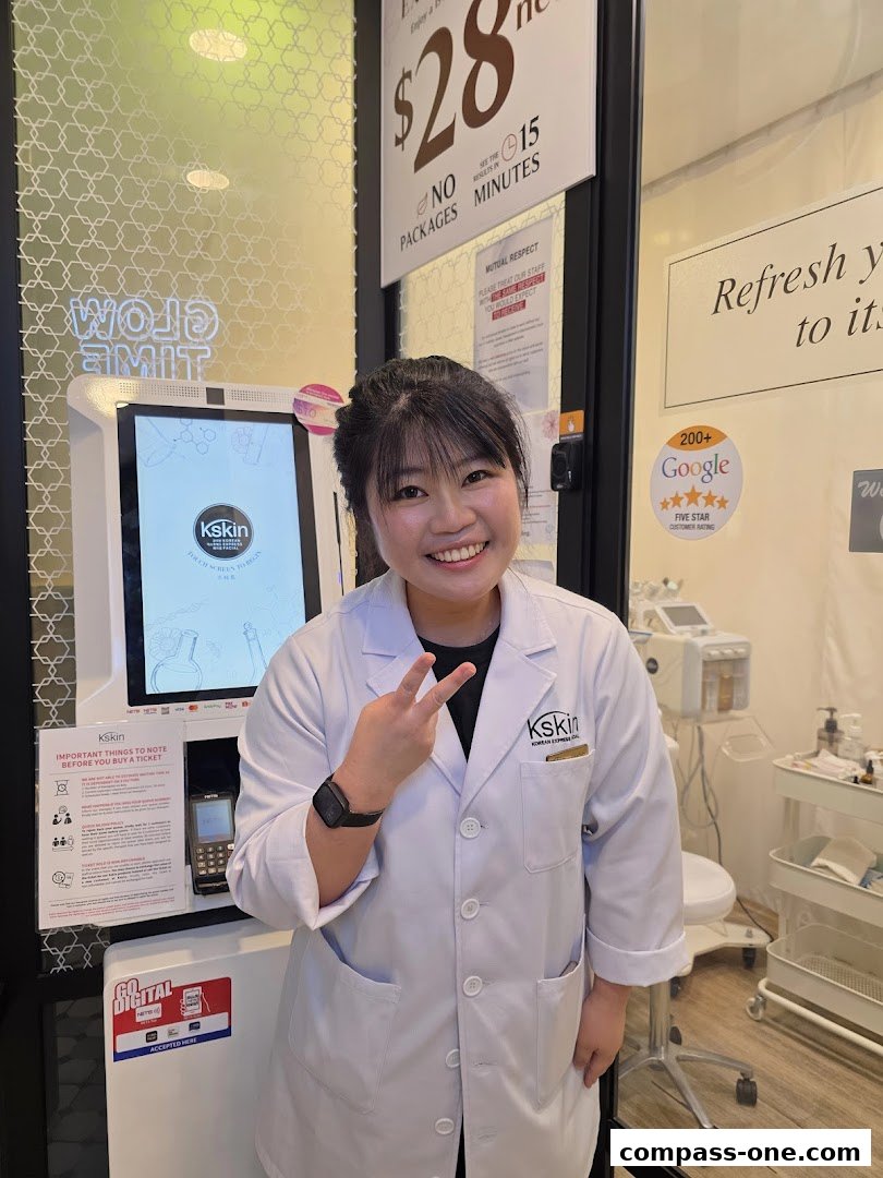A smiling employee in a white lab coat poses in front of a Kskin Korean Express Facial kiosk.