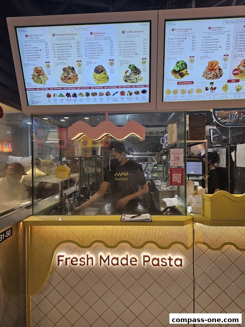 A brightly lit pasta shop counter with a menu board displaying various pasta dishes and a staff member preparing food.