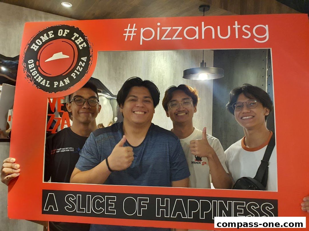 Four young men pose for a photo in a Pizza Hut branded frame, smiling and giving thumbs up.