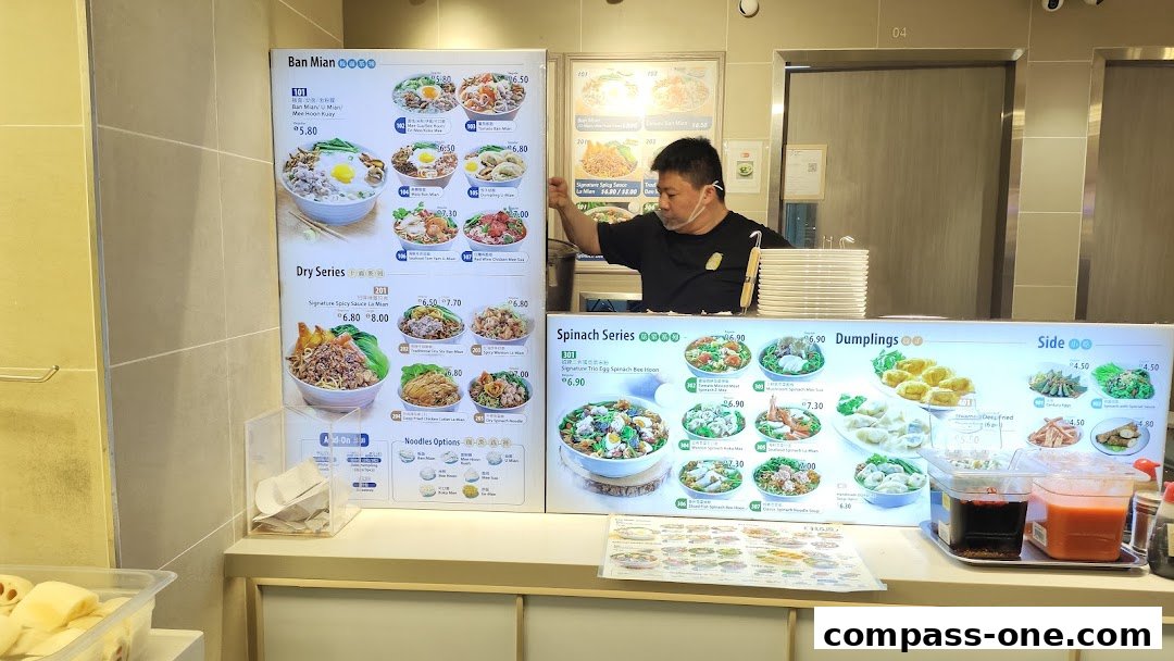 A food stall counter displaying a menu with various noodle dishes, dumplings, and sides.
