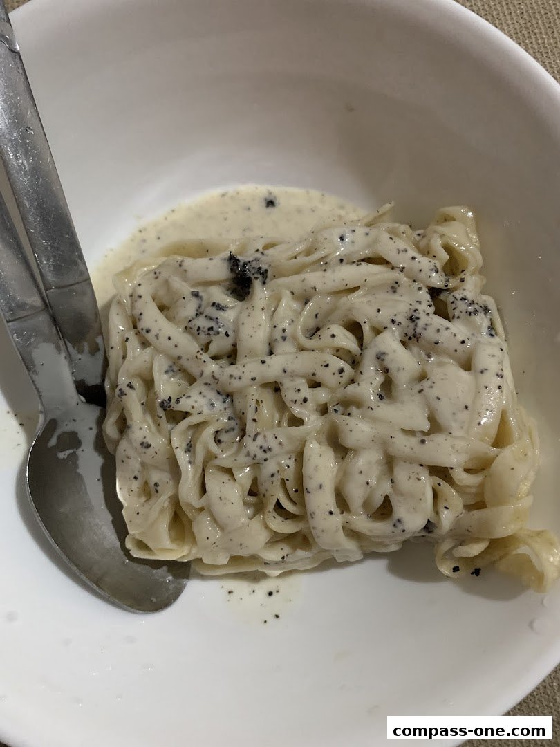 A close-up shot of fresh pasta with a creamy sauce and black specks, served in a white bowl with cutlery.