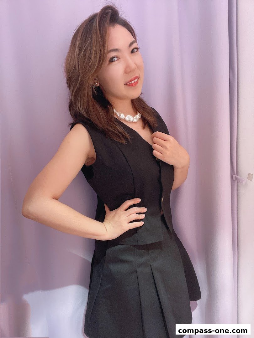 A woman in a black outfit and pearl necklace poses in front of a purple curtain.