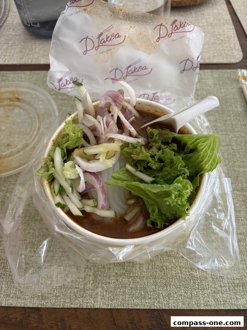 A bowl of Laksa with fresh vegetables and a white spoon, branded packaging in the background.